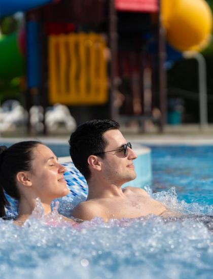 Coppia rilassata in piscina con scivolo colorato sullo sfondo.