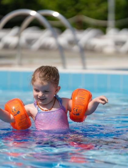 Bambina con braccioli arancioni gioca in piscina con un adulto.