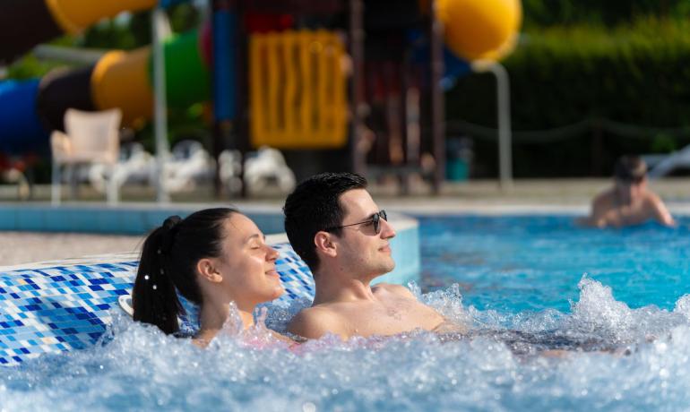 Coppia rilassata in piscina con scivolo colorato sullo sfondo.