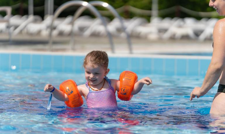 Bambina con braccioli arancioni gioca in piscina con un adulto.