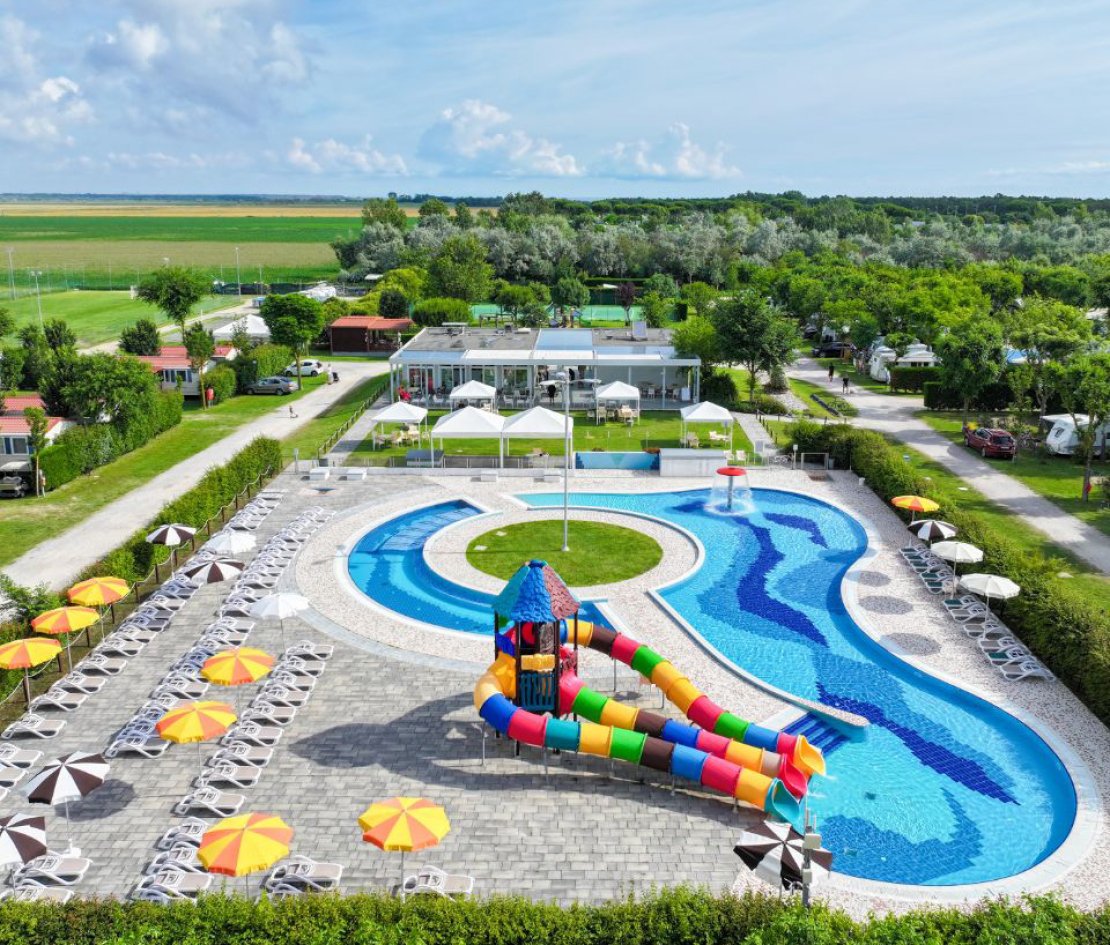 Piscina con scivoli colorati e ombrelloni, circondata da verde e strutture ricreative.