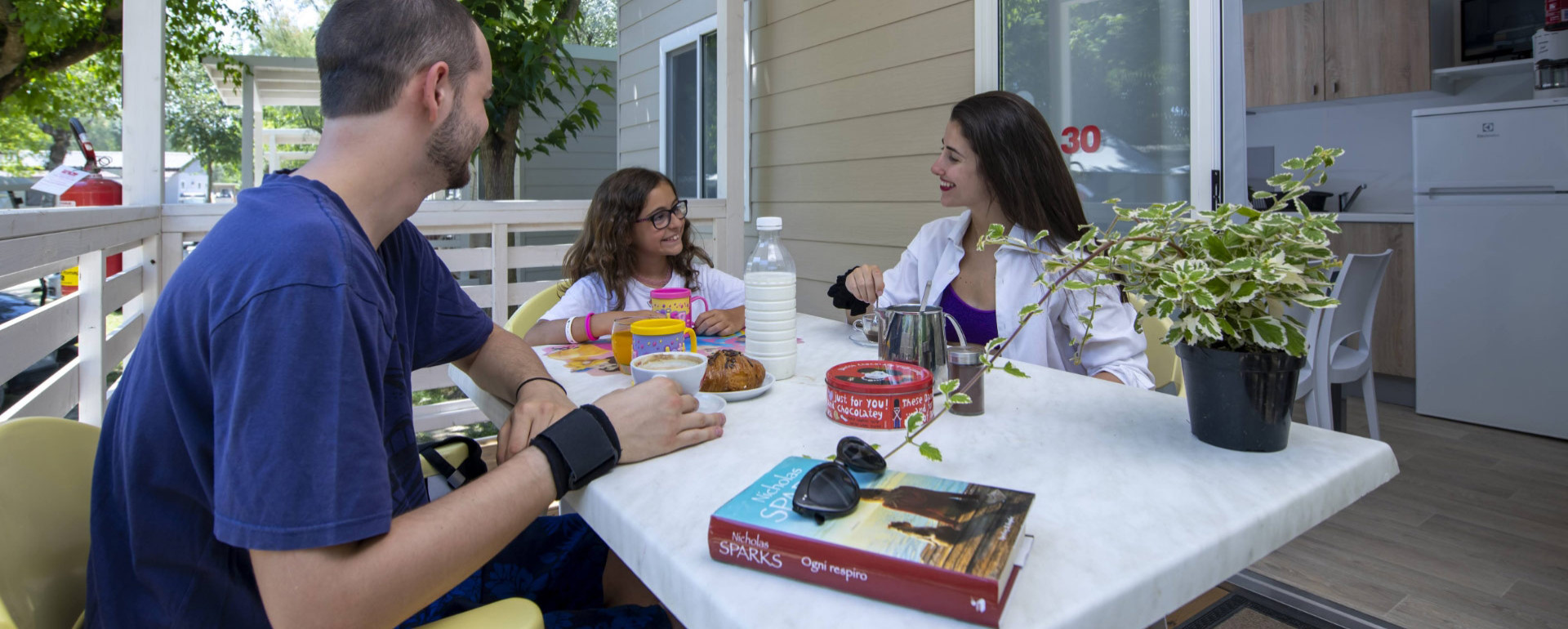 Famiglia fa colazione all'aperto su un terrazzo, con libro e piante sul tavolo.