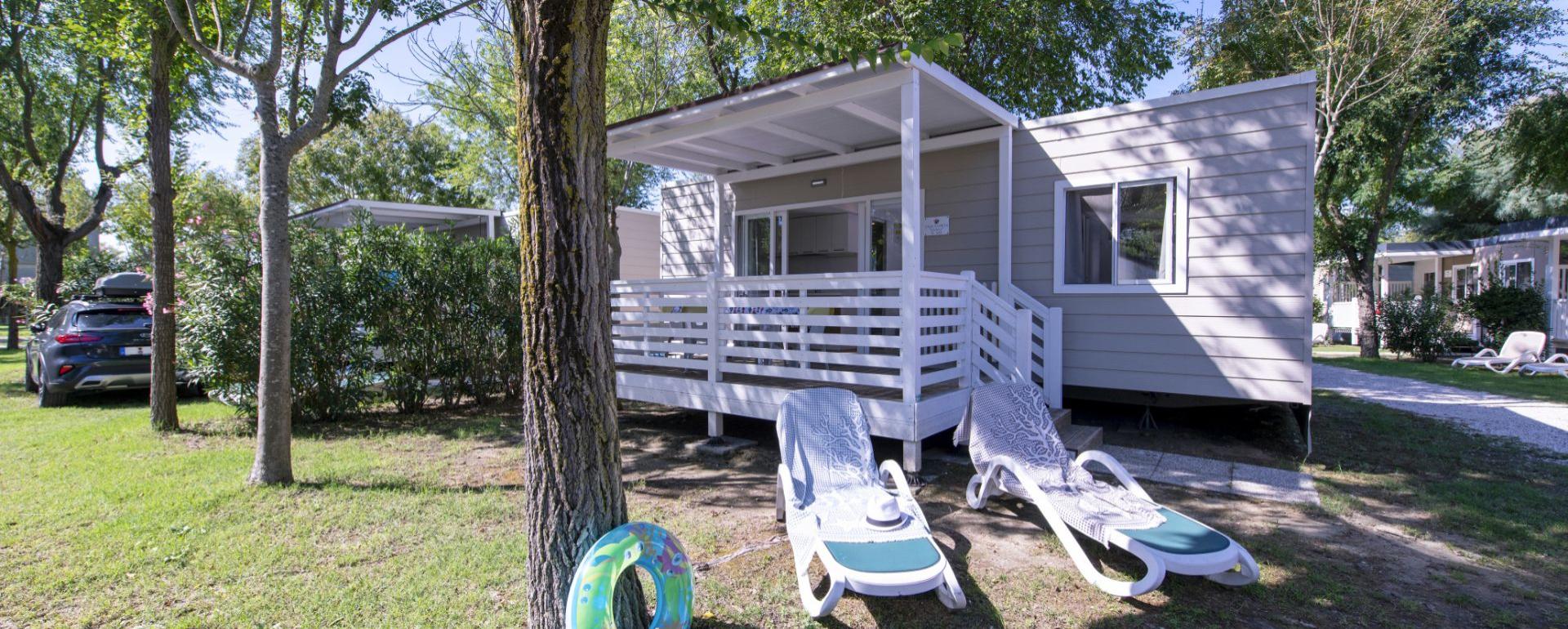 Bungalow met veranda en ligstoelen in een met bomen omzoomde camping.