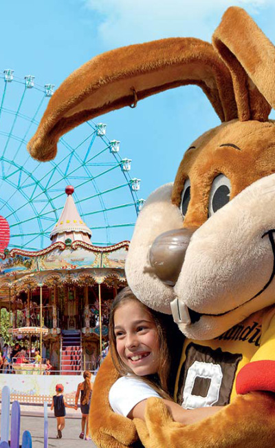 Parco divertimenti con montagne russe, ruota panoramica e mascotte che abbraccia una bambina sorridente.