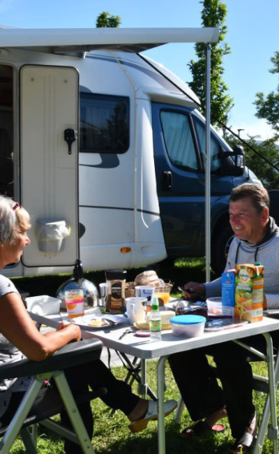 Coppia fa colazione all'aperto accanto a un camper, moto e bici parcheggiate.