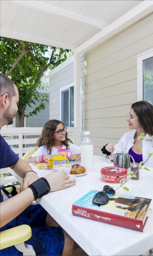 Famiglia fa colazione all'aperto, libri e dolci sul tavolo, atmosfera serena.