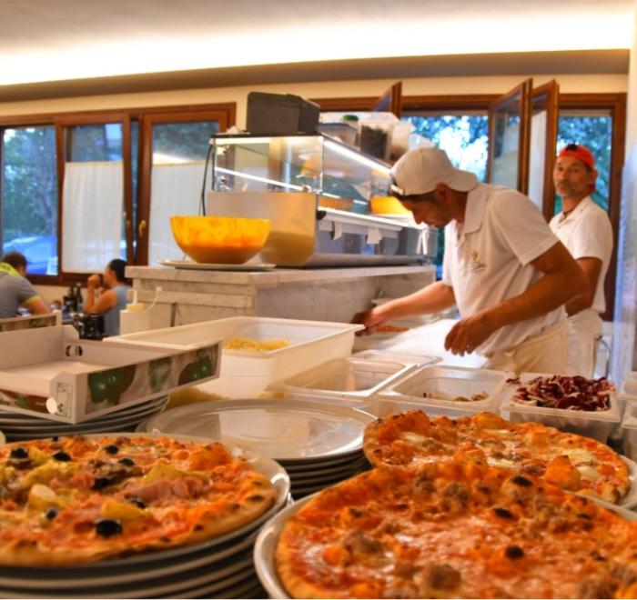 Pizzaioli preparano pizze in una pizzeria affollata con ingredienti freschi.