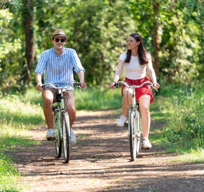 Due persone in bicicletta su un sentiero nel bosco, sorridono e si godono la natura.