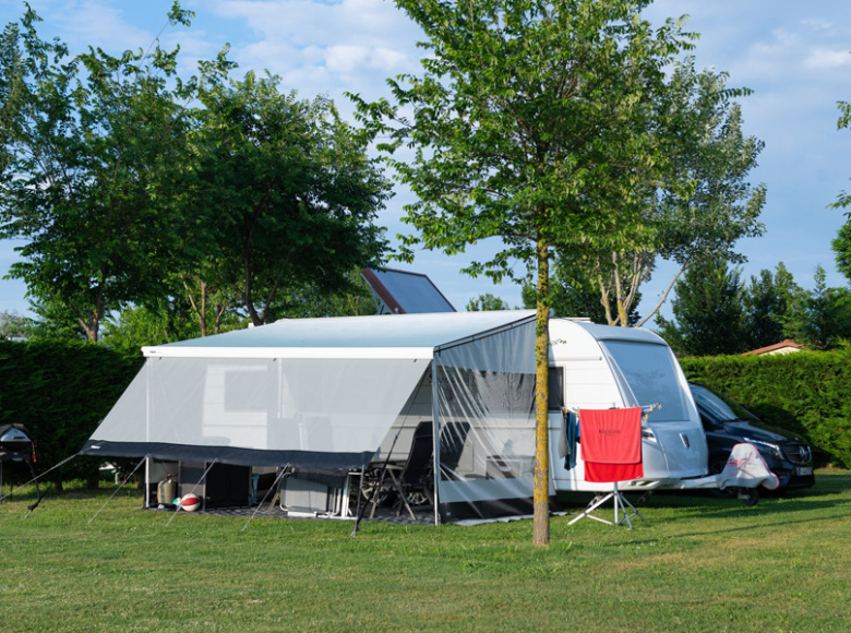 Camper con veranda in campeggio, circondato da alberi e prato verde.