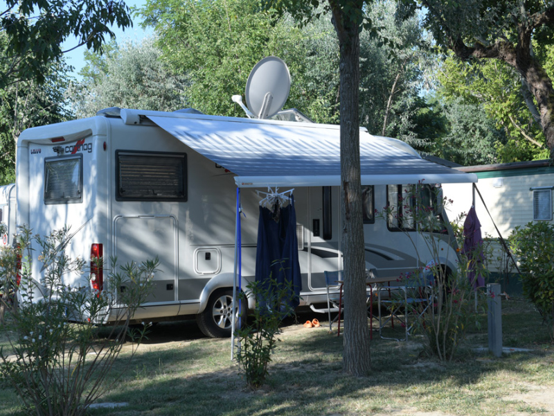 Camper parcheggiato in campeggio con veranda aperta e vegetazione intorno.