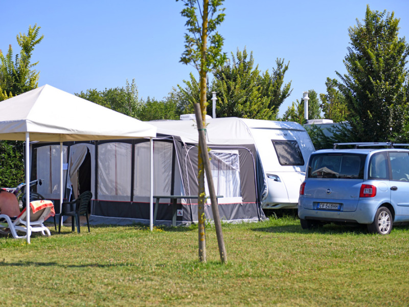 Campeggio con roulotte, tenda, auto e biciclette su prato verde, giornata soleggiata.